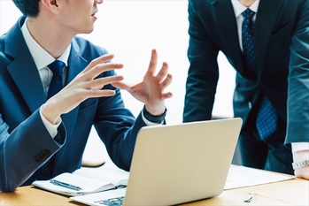 デスクに座りノートパソコンを前に同僚や上司とビジネスの話をする2人のスーツ姿の顔のない男性